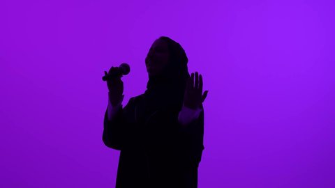 بدء التدريب الصوتي، قضاء وقت ممتع بأداء العروض الصوتية، تصوير ظلي مقرب لشابة عربية خليجية سعودية محجبة ترتدي العباءة تمسك المايكروفون، سيدة عمانية تمارس موهبة الغناء بإيماءات السرور، خلفيه بنفسجية، سالوييت