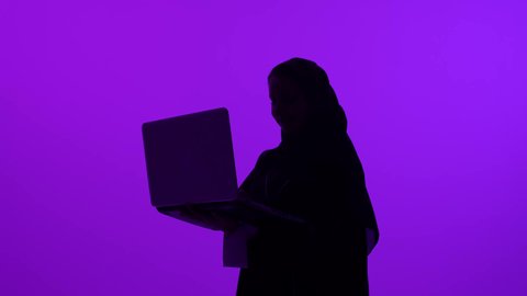 Using a modern and advanced technical device, managing work remotely, close-up silhouette of a veiled Omani Gulf Arab woman wearing an abaya working on a laptop, a Saudi woman completing required tasks via a laptop, purple background, silhouette