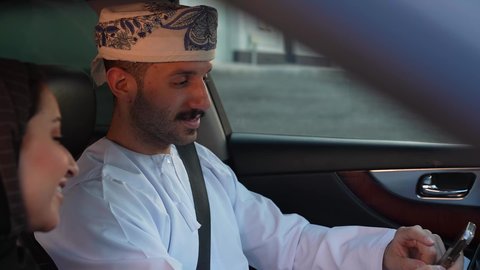 Walking in the streets of the Sultanate of Oman with the family, a family group trip in the car, chatting and talking on the road, an Arab Gulf Omani man wearing a dishdasha and turban sitting with his wife inside the car, taking a recreational tour with gestures of happiness and pleasure, spending a fun time with the family, wearing a seat belt