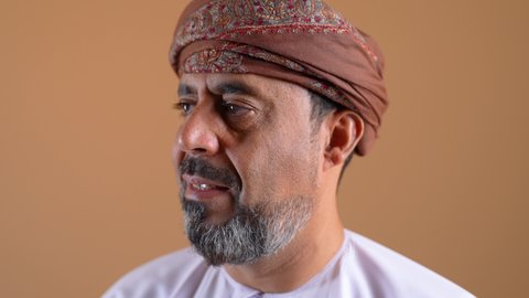 الاناقة والاهتمام بالمظهر الخارجي، تعابير الثقة بالنفس، مفهوم الرجولة والوقار، تصوير مقرب لرجل عربي خليجي عماني يرتدي الدشداشة والعمامة يقف باستقامة ينظر الى الكاميرا ما بايماءات السرور، خلفية بيج