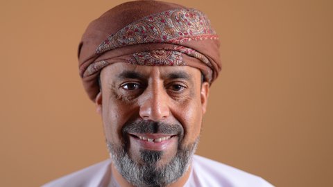 Expressions of self-confidence, the concept of masculinity and dignity, elegance and interest in outward appearance, a close-up of an Arab Gulf Omani man wearing a dishdasha and turban, standing straight, looking at the camera with gestures of pleasure, beige background