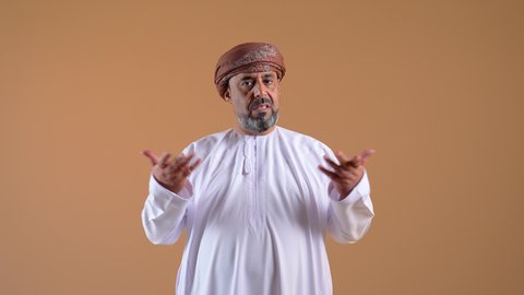 التردد بشأن أمر معين، عدم التاكد والقدرة على اتخاذ القرار، تصوير مقرب لرجل عربي خليجي عماني يرتدي الدشداشة والعمامة يرفع كفيه بتعابير الحيرة، النظر إلى مكان ما بإيماءات الاستغراب، خلفية بيج