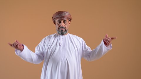 Locating something, hand gestures indicating this place (here), close-up of a smiling Omani Gulf Arab man wearing a dishdasha and turban pointing his index fingers down, beige background