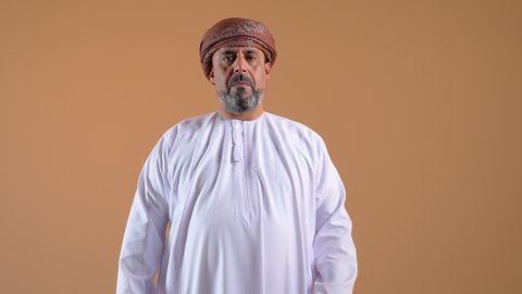 Hesitation about a certain matter, lack of certainty and inability to make a decision, close-up of an Arab Gulf Omani man wearing a dishdasha and turban raising his hands with an expression of confusion, looking somewhere with gestures of surprise, beige background