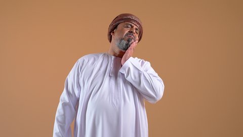 مفهوم تسوس الأضراس, تصوير مقرب لرجل عربي خليجي عماني يرتدي الدشداشة والعمامة يضع يده على وجهه بإيماءات الألم و وجع الأسنان، الإنتفاخ والإلتهاب بالفم واللثة، ضرورة الذهاب للطبيب والحصول على العلاج، خلفية بيج