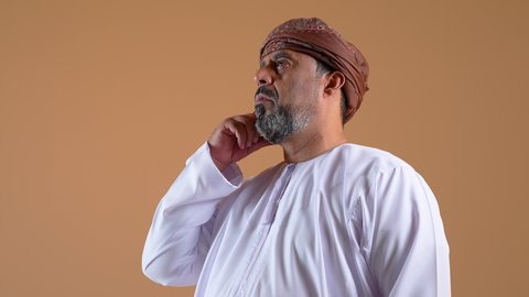 تعابير شرود الذهن والتمعن، التأمل والتخطيط للمستقبل، ايجاد حل لمشكلة ما، تصوير مقرب لرجل عربي خليجي عماني يرتدي الدشداشة والعمامة يضع يده على رأسه بايماءات التفكير والتركيز، المشاكل والهموم والضغوطات المحيطة، خلفية بيج