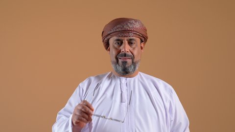 مفهوم الرجولة والوقار، تعابير الثقة بالنفس، الاهتمام بالمظهر الخارجي، تصوير مقرب لرجل عربي خليجي عماني يرتدي الدشداشة والعمامة يضع النظارة الشمسية ينظر إلى الاعلى بايماءات التركيز، خلفية بيج