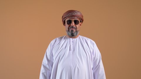 Expressions of self-confidence, the concept of masculinity and dignity, elegance and interest in outward appearance, a close-up of an Arab Gulf Omani man wearing a dishdasha and turban, standing straight, looking at the camera with gestures of pleasure, beige background