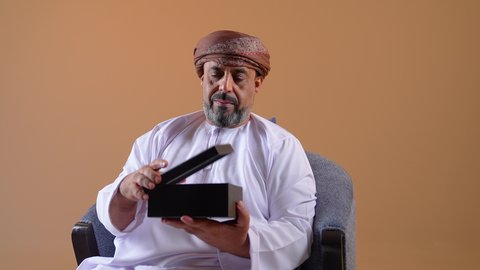 مفهوم الشكر والامتنان، تقديم الهدايا بالاعياد والمناسبات الخاصة، التعبير عن الحب والتقدير والاحترام، تصوير مقرب لرجل عربي خليجي عماني يرتدي الدشداشة والعمامة يجلس على كرسي مريح يحمل بيديه صندوق هدية, ايماءات المفاجئة والصدمة، خلفية بيج