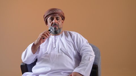 الدورات التدريبية و ورشات العمل، إلقاء المحاضرات وإقامة الندوات، تبادل الحوار والاستفادة من خبرات الكبار، تصوير مقرب لرجل عربي خليجي عماني يرتدي الدشداشة والعمامة يجلس على كرسي مريح يحمل بيده مكبر الصوت، خلفية بيج