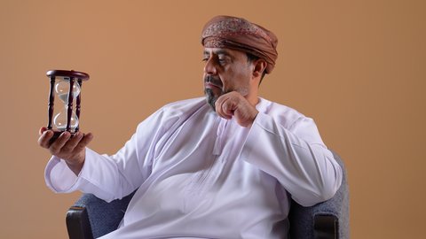 القيام بمراقبة توقيت الرمل, إدراك قيمة الوقت, مفهوم إدارة الأعمال وتنظيم الجدول الزمني, تصوير مقرب لرجل عربي خليجي عماني يرتدي الدشداشة والعمامة يجلس على أريكة مريحة يحمل بيده ساعة رملية زجاجية (البنكام)، خلفية بيج