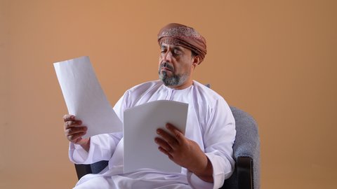 مراجعة ومتابعة الخطط والمشاريع المكتبية، اعداد التقارير الخاصة بالعمل، تصوير مقرب لرجل عربي خليجي عماني يرتدي الدشداشة والعمامة يحمل بيده مجموعة من الاوراق، التدقيق في قراءة الملاحظات، خلفية بيج