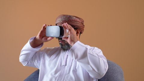 Merging modern technology and technology with fun and entertainment, simulating the imaginary world, close-up of an Arab Gulf Omani man wearing a dishdasha, turban and 3D virtual reality glasses, gestures of integration and focus, beige background