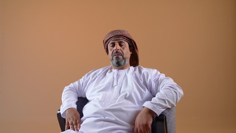 إيماءات الراحة والإسترخاء، تصوير مقرب لرجل عربي خليجي عماني يرتدي الدشداشة والعمامة يجلس على كرسي مريح يضع يديه اسفل رأسه يغمض عينيه بتعابير النعاس، الشعور بالتعب والارهاق، خلفية بيج