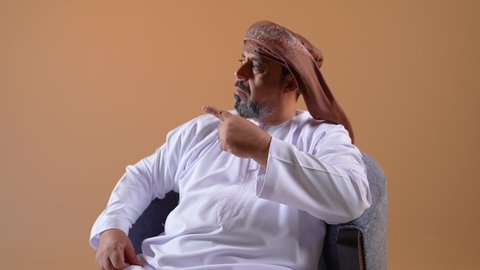 حركات يدين تدل على هذا المكان (هنا)، تحديد موقع شيء ما، بورتريه لرجل عربي خليجي عماني مبتسم يرتدي الدشداشة والعمامة يجلس على أريكة مريحة يشير بأصبع السبابة نحو الاعلى بايماءات السرور والسعادة، خلفية بيج