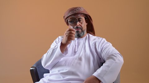 مفهوم الفحص والاستكشاف، توضيح الصورة والرؤية عن قرب، تصوير مقرب لرجل عربي خليجي عماني يرتدي الدشداشة والعمامة يجلس على كرسي مريح يحمل بيده عدسة مكبرة يضعها على عينه بإيماءات التركيز والتمعن، خلفية بيج