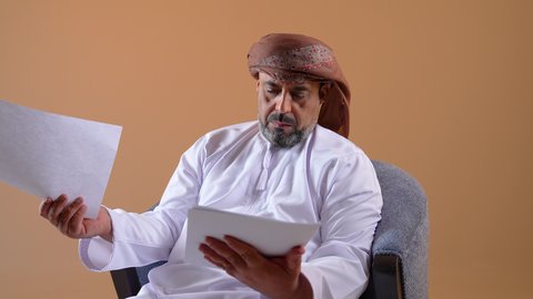 مراجعة ومتابعة الخطط والمشاريع المكتبية، اعداد التقارير الخاصة بالعمل، تصوير مقرب لرجل عربي خليجي عماني يرتدي الدشداشة والعمامة يحمل بيده مجموعة من الاوراق، التدقيق في قراءة الملاحظات، خلفية بيج