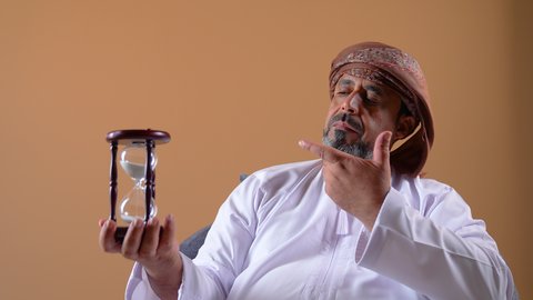القيام بمراقبة توقيت الرمل, إدراك قيمة الوقت, مفهوم إدارة الأعمال وتنظيم الجدول الزمني, تصوير مقرب لرجل عربي خليجي عماني يرتدي الدشداشة والعمامة يجلس على أريكة مريحة يحمل بيده ساعة رملية زجاجية (البنكام)، خلفية بيج