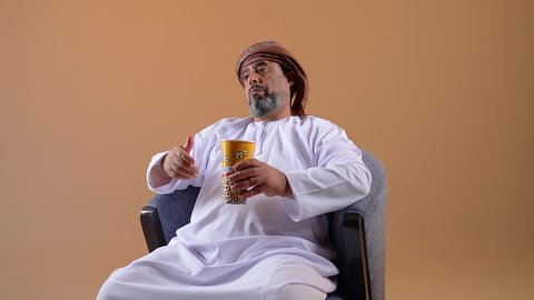 Building and strengthening body muscles, lifting heavy iron weights, close-up of an Arab Gulf Omani man wearing a dishdasha and turban sitting on a comfortable chair holding weights in his hand, physical activity to maintain physical fitness, beige background