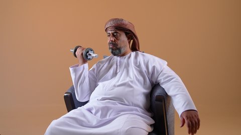 بناء عضلات الجسم وتقويتها، رفع الأوزان الحديدية الثقيلة، تصوير مقرب لرجل عربي خليجي عماني يرتدي الدشداشة والعمامة يجلس على كرسي مريح يحمل بيده الأثقال، نشاط بدني للمحافظة على اللياقة الجسدية، خلفية بيج