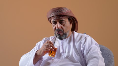 Trying to relieve the summer heat with refreshing juices, enjoying a delicious and healthy drink, a close-up of an Omani Gulf Arab man wearing a dishdasha and turban sitting on a comfortable chair drinking cold natural juice, beige background