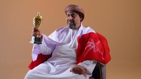 التتويج بكأس البطولة بعد تحقيق الانتصار، تشجيع المنتخب الوطني العماني، تصوير مقرب لرجل عربي خليجي عماني يرتدي الدشداشة العمامة يجلس على كرسي مريح يضع علم سلطنة عمان على كتفيه، خلفية بيج