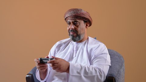 Having fun at home, home entertainment activity, using modern technology in entertainment, close-up of an Arab Gulf Omani man sitting on a comfortable chair holding a joystick in his hand playing video games with gestures of happiness and pleasure, fun, entertainment and interaction, beige background