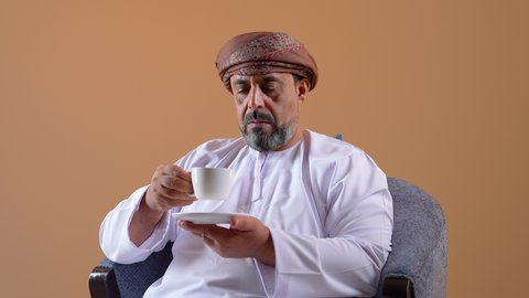 الاستمتاع بالمشروبات الساخنة، تناول الأعشاب الشتوية الدافئة، قضاء وقت الفراغ باحتساء المشروب المفضل, تصوير مقرب لرجل عربي خليجي عماني مبتسم يرتدي الدشداشة والعمامة يجلس على كرسي مريح يقوم بشرب القهوة، خلفية بيج