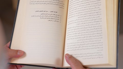 Spending free time learning and culture, interest in browsing useful books, the concept of the love of reading, a close-up photo of a forty-year-old Arab Gulf Omani man wearing a dishdasha and turban sitting on a comfortable chair holding a book in his hand, beige background