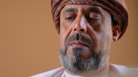 مفهوم حب المطالعة والقراءة، تصوير مقرب لرجل اربعيني عربي خليجي عماني  يرتدي الدشداشه والعمامة يجلس على كرسي مريح يحمل بيده كتاب، قضاء وقت الفراغ بالتعلم والتثقف، الاهتمام بتصفح الكتب المفيدة، خلفية بيج