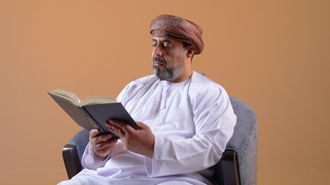 الاهتمام بتصفح الكتب المفيدة، مفهوم حب المطالعة والقراءة، تصوير مقرب لرجل اربعيني عربي خليجي عماني  يرتدي الدشداشه والعمامة يجلس على كرسي مريح يحمل بيده كتاب، قضاء وقت الفراغ بالتعلم والتثقف، خلفية بيج