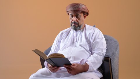مفهوم حب المطالعة والقراءة، تصوير مقرب لرجل اربعيني عربي خليجي عماني  يرتدي الدشداشه والعمامة يجلس على كرسي مريح يحمل بيده كتاب، قضاء وقت الفراغ بالتعلم والتثقف، الاهتمام بتصفح الكتب المفيدة، خلفية بيج