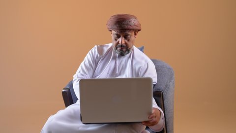 عقد إجتماع عمل عبر الإنترنت، مناقشة إستراتيجية المشروع, إدارة شؤون الأعمال وتنظيمها، استخدام جهاز تقني حديث ومتطور، تصوير مقرب لرجل عربي خليجي عماني يرتدي الدشداشة والعمامة يجلس على كرسي مريح يقوم بإجراء مكالمة فيديو من خلال الحاسب المحمول, خلفية بيج