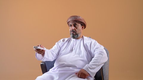 Changing and flipping between channels, the concept of guidance and control, the use of modern technical devices, a close-up of an Arab Gulf Omani man wearing a dishdasha and turban sitting on a comfortable chair holding a remote control in his hand, watching movies and entertainment programs, beige background