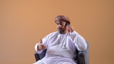 استخدام جهاز تقني حديث ومتطور، اجراء مكالمة هاتفية عبر الجوال، التواصل مع الأهل والأصدقاء، الاطمئنان على الأقارب وسماع اخبارهم، تصوير مقرب لرجل عربي خليجي عماني يرتدي الدشداشة والعمامة يجلس على كرسي مريح يحمل بيده الهاتف المحمول، خلفية بيج