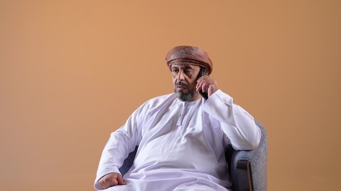 Making a phone call via mobile phone, communicating with family and friends, using a modern and advanced technical device, checking on relatives and hearing their news, a close-up of an Arab Gulf Omani man wearing a dishdasha and turban sitting on a comfortable chair holding a mobile phone in his hand, beige background