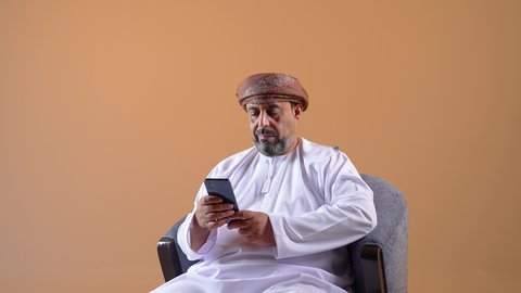 تصفح مواقع التواصل الاجتماعي، التواصل مع الأهل والأصدقاء، استخدام جهاز تقني حديث ومتطور، الاطمئنان على الأقارب وسماع اخبارهم، تصوير مقرب لرجل عربي خليجي عماني يرتدي الدشداشة والعمامة يجلس على كرسي مريح يحمل بيده الهاتف المحمول، خلفية بيج
