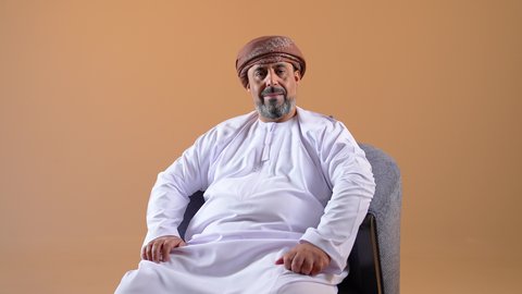 الاهتمام بالمظهر الخارجي، مفهوم الرجولة والوقار، ايماءت الثقة بالنفس، تصوير مقرب لرجل عربي خليجي عماني يرتدي الدشداشة والعمامة يجلس على كرسي مريح ينظر الى الكاميرا بتعابيرالسعادة والسرور، خلفية بيج