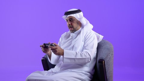 قضاء وقت ممتع ومسلي، رجل عربي خليجي كويتي يرتدي الثوب التقليدي يجلس على كرسي مريح يمسك بيديه عصا التحكم، التفاعل والاندماج مع ألعاب الفيديو، رجل سعودي يستخدم الأجهزة والتقنيات الإلكترونية الحديثة