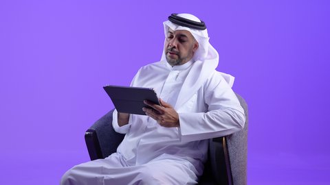 استخدام الأجهزة والتقنيات الحديثة المتطورة، اجتماع عمل عبر الإنترنت، رجل عربي خليجي كويتي يرتدي الثوب التقليدي يجلس على كرسي مريح يقوم بإجراء مكالمة فيديو عبر جهاز التابلت ، رجل سعودي يدير شؤون الأعمال وينظمها عن بعد
