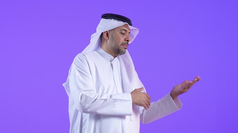 النظر إلى الكاميرا بإيماءات الفرح والسرور، رجل عربي خليجي كويتي مبتسم يرتدي الثوب التقليدي والغترة يقف ويشير بيديه جانبا إلى شيء محدد بتعابير هذا المكان هنا، رجل سعودي يحدد موقع معين