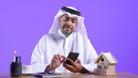 دمج التقنيات الحديثة بالمجال الإداري والمكتبي، الاستمتاع بتناول فنجان قهوة ساخنة، رجل عربي خليجي كويتي يرتدي الثوب التقليدي والغترة يجلس على مكتب بجانبه بعض الاقلام ويمسك الهاتف المحمول ،موظف سعودي يستخدم التقنيات الحديثة في الأعمال الإدارية والمكتبية