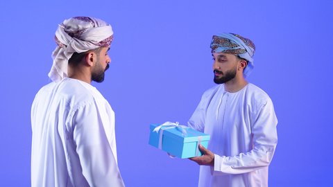 التعبير عن المحبة والألفة بين الاصدقاء، تقديم الهدايا التذكارية، أعياد ومناسبات ومفاجآت سعيدة, بورتريه مقرب لشاب عربي خليجي عماني يرتدي الدشداشة و العمامة يقوم بتقديم الهدية لصديقة, خلفية زرقاء