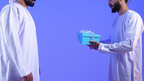 تقديم الهدايا التذكارية، أعياد ومناسبات ومفاجآت سعيدة, التعبير عن المحبة والألفة بين الاصدقاء, بورتريه مقرب لشاب عربي خليجي عماني يرتدي الدشداشة و العمامة يقوم بتقديم الهدية لصديقة, خلفية زرقاء