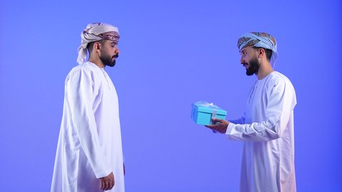 أعياد ومناسبات ومفاجآت سعيدة, تقديم الهدايا التذكارية, التعبير عن المحبة والألفة بين الاصدقاء, بورتريه لشاب عربي خليجي عماني يرتدي الدشداشة و العمامة يقوم بتقديم الهدية لصديقة, خلفية زرقاء
