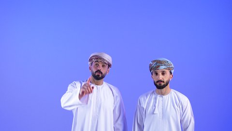 مفهوم الأناقة والاهتمام بالمظهر الخارجى، الإشارة إلى شيء ما، النظر إلى الأعلى بإيماءات السعادة و السرور, بورترية مقرب لرجلان عربيان خليجيان عمانيان يرتديان الدشداشة و العمامه يشيران بإصبع السبابة نحو شيء معين, خلفية زرقاء