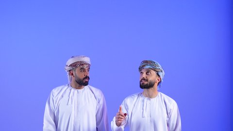 Pointing at something, looking up with gestures of happiness and pleasure, close-up portrait of two Omani Gulf Arab men wearing dishdasha and turban pointing with index finger towards something, blue background