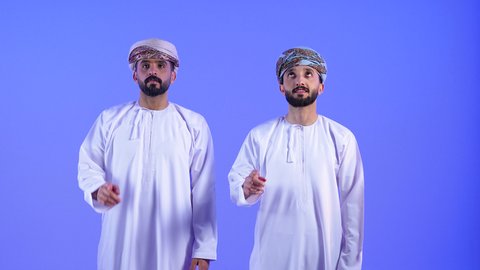 Looking up with gestures of happiness and pleasure, pointing at something, portrait of two Omani Gulf Arab men wearing dishdasha and turban pointing with index finger towards something, blue background