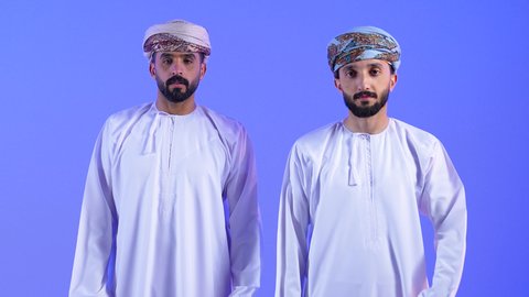 تحديد موقع شيء معين، إشارات وحركات يدين تدل على هذا المكان (هنا), بورتريه لشابان عربيان خليجيان عمانيان يرتديان االدشداشة و العمامة يقومان بالاشارة بإصبع السبابة إلى شيء محدد, خلفية زرقاء
