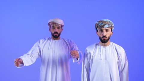 الوقوع في مصيبة, الانزعاج عند سماع خبر سيء , النظر الى الكاميرا بتعابير التعجب, الصدمة و المفاجأة برؤية شيء ما، بورتريه لرجلان عربيان خليجيان عمانيان يرتديان الدشداشة و العمامه   يضعان يدهما على وجههم بتعابير الدهشة و الذهول, خلفية زرقاء